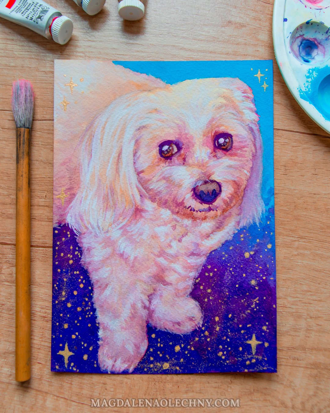 A painting of a Maltese dog on a galaxy background. The portrait is surrounded by paints, a brush, and a palette.