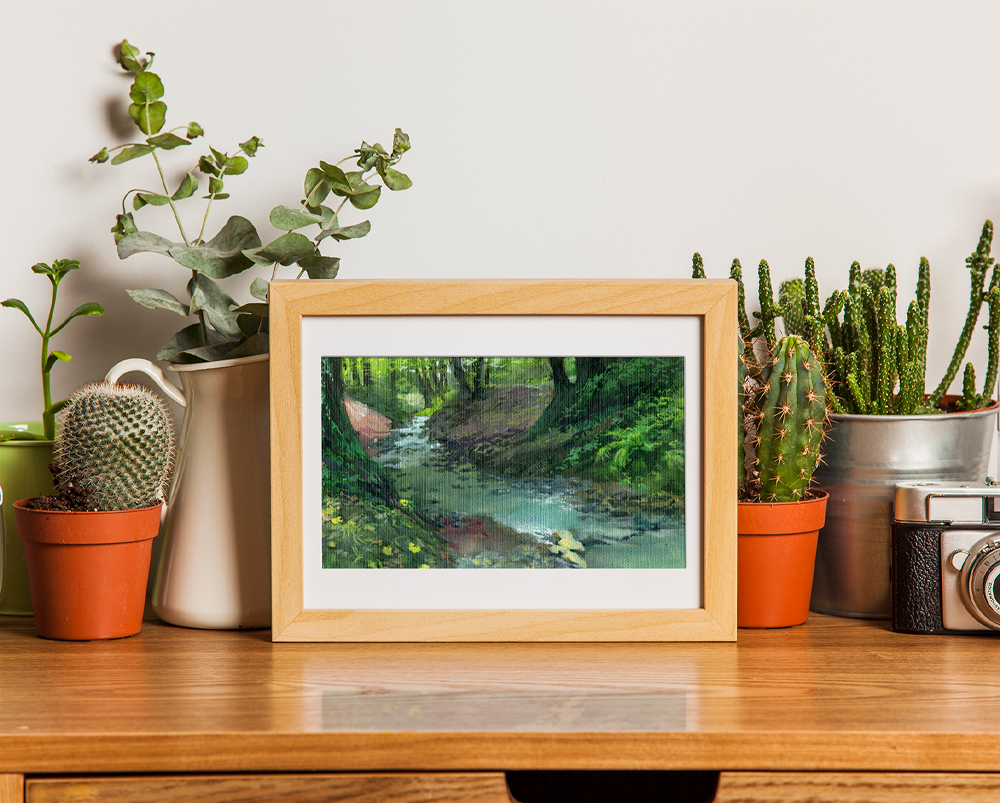 Framed gouache painting of a stream in the park. The stream is rather shallow and narrow, surrounded by trees and muddy banks covered by greenery. The weather is overcast. There are plants around the painting.