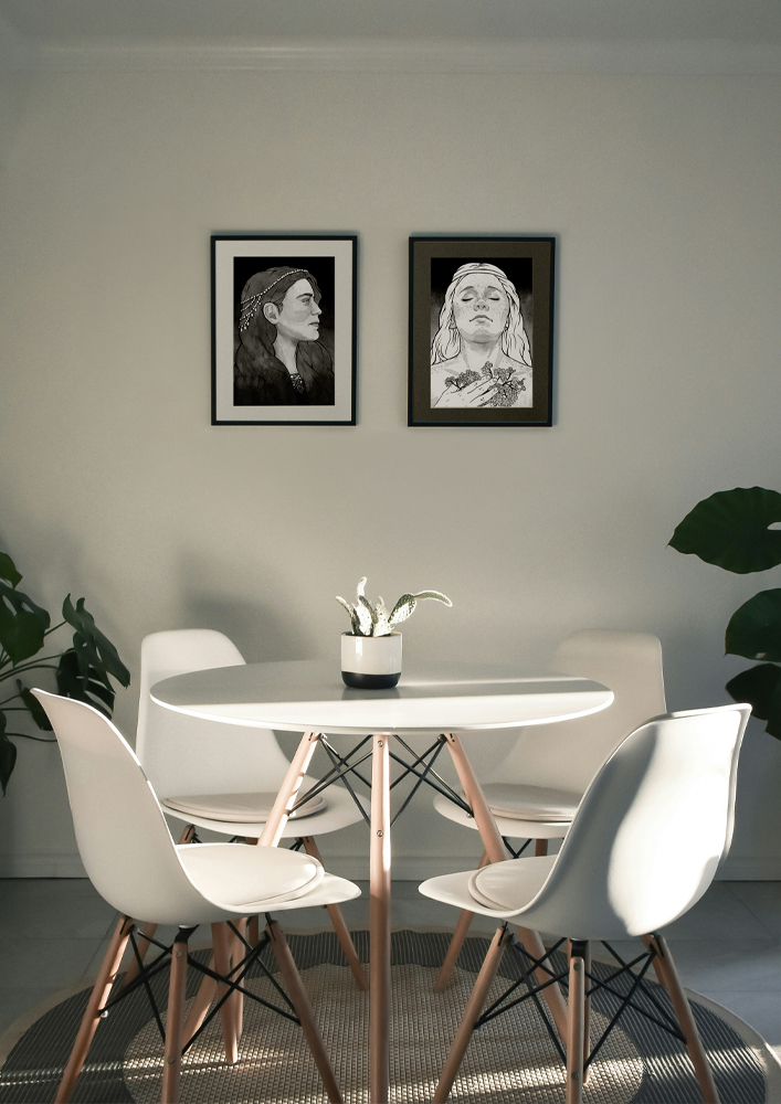 A room with two framed paintings, "Dark Maiden, Noble and Proud" and "Daughter of Tears" by artist Magdalena Olechny. They hang above a table with four chairs.