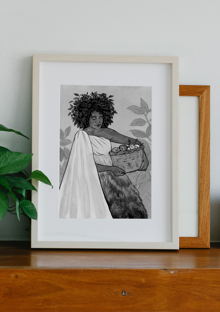 Framed original painting inspired by Yavanna from “The Silmarillion” by J.R.R. Tolkien. Ink drawing of a black woman with leaf hair in a dress adorned with leafy branches. She is carrying a basket full of fruit, vegetables, and mushrooms. Behind the painting, there is another frame and next to it there is a plant.