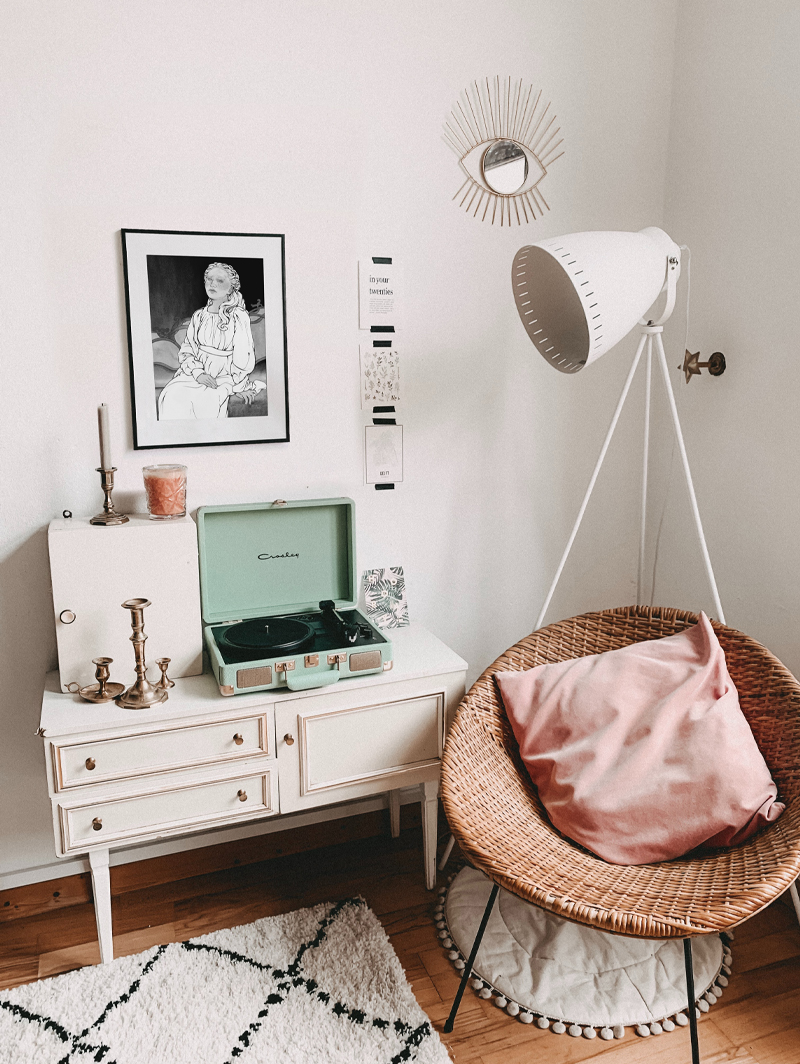 A photo of a cozy room with a rattan chair and a pillow, two rugs, a standing lamp, a dresser, and a framed Finduilas print. There are a few candle holders, candles, and a record player on the dresser.