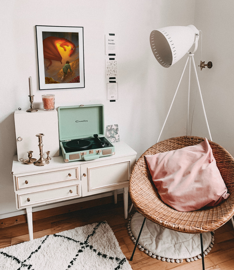 A photo of a cozy room with a rattan chair and a pillow, two rugs, a standing lamp, a dresser, and a framed Hobbit print. There are a few candle holders, candles, and a record player on the dresser.