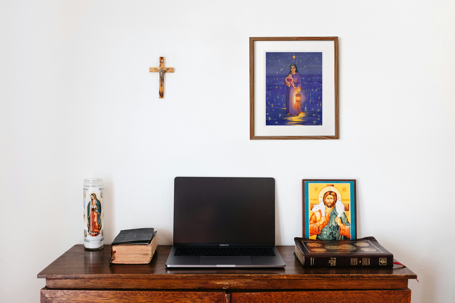 A photo of a room. There is a desk with a laptop, the Bible, Jesus' icon, an old book, a candle with Our Lady of Guadalupe, and a framed print above it. A print depicts a painting of Stella Maris, Our Lady, Star of the Sea.