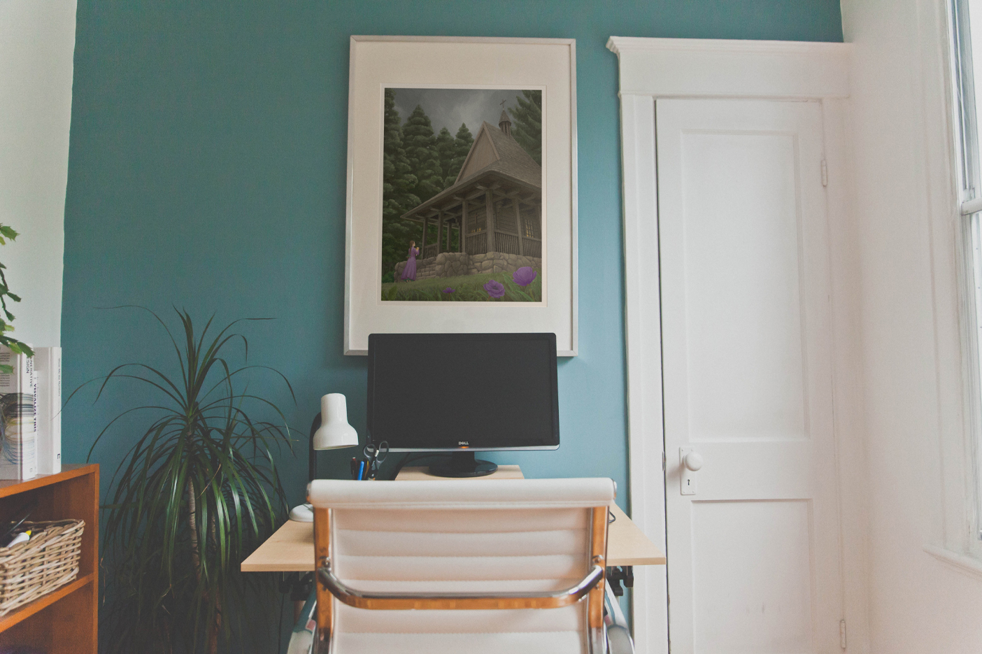 A photo of a living room. There is a desk with a screen, a lamp next to it, a chair in front of the desk, and a framed print above it. There are doors to the right, a plant, and a rack to the left of the desk.