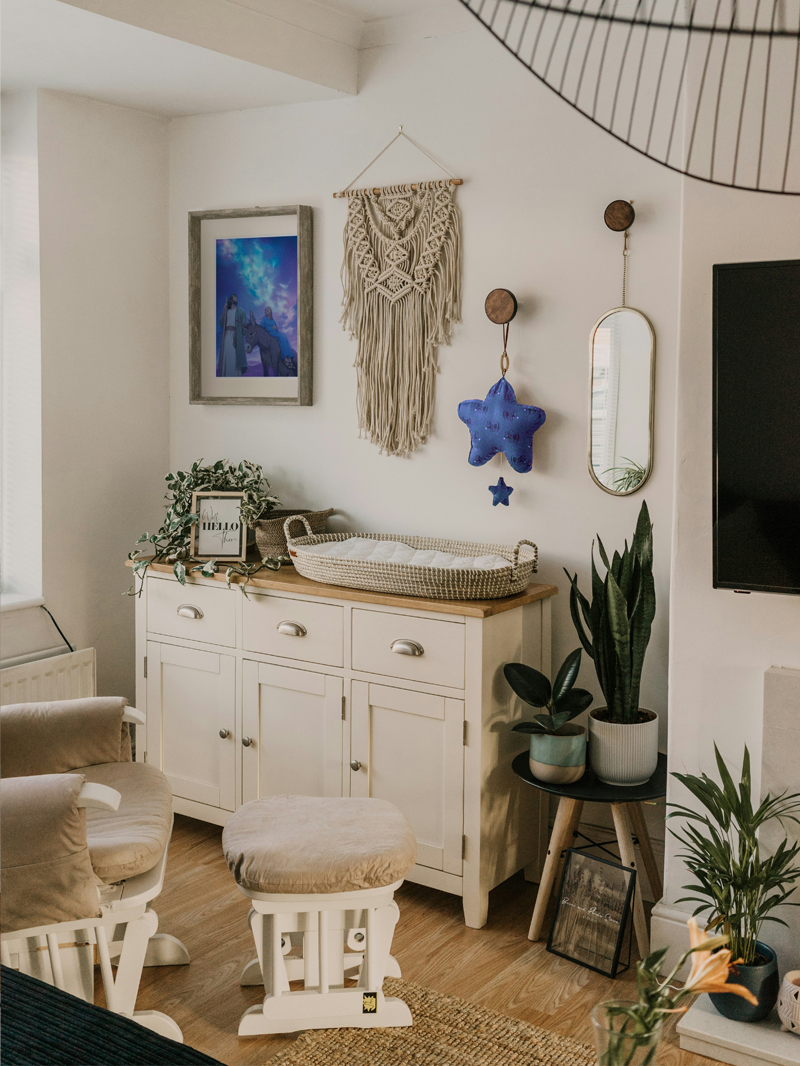 A photo of a nursery room. There is a place to change the baby, with a framed print, a macrame and plush stars above it, and an armchair with a footrest. The room is filled with plants.