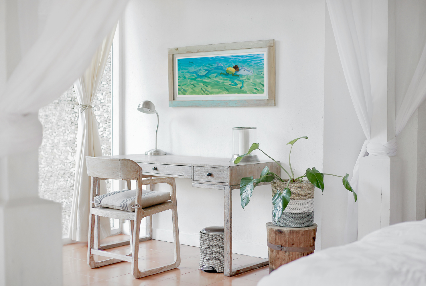 A photo of a sleeping room. There is a desk with a lamp, a plant next to it, and a framed print above it. We can see a fragment of the bed in the foreground.